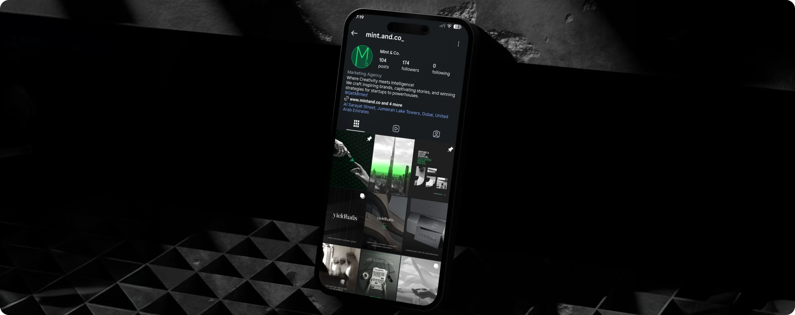 Phone showing the Mint & Co. Instagram profile on a dark geometric surface